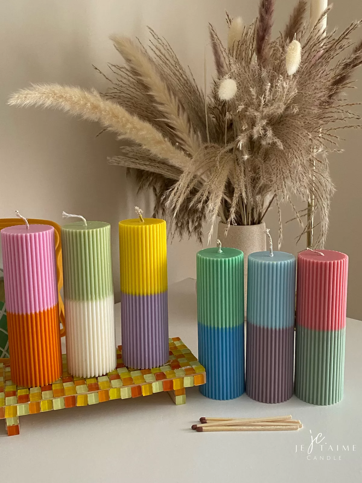 A row of tall ribbed pillar candles in pastel and bright two-tone colors, arranged on a white surface. The candles are displayed beside a green checkered lamp, a decorative mirror with an orange frame, and pampas grass in the background.