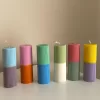 A row of tall ribbed pillar candles in pastel and bright two-tone colors, arranged on a white surface. The candles are displayed beside a green checkered lamp, a decorative mirror with an orange frame, and pampas grass in the background.