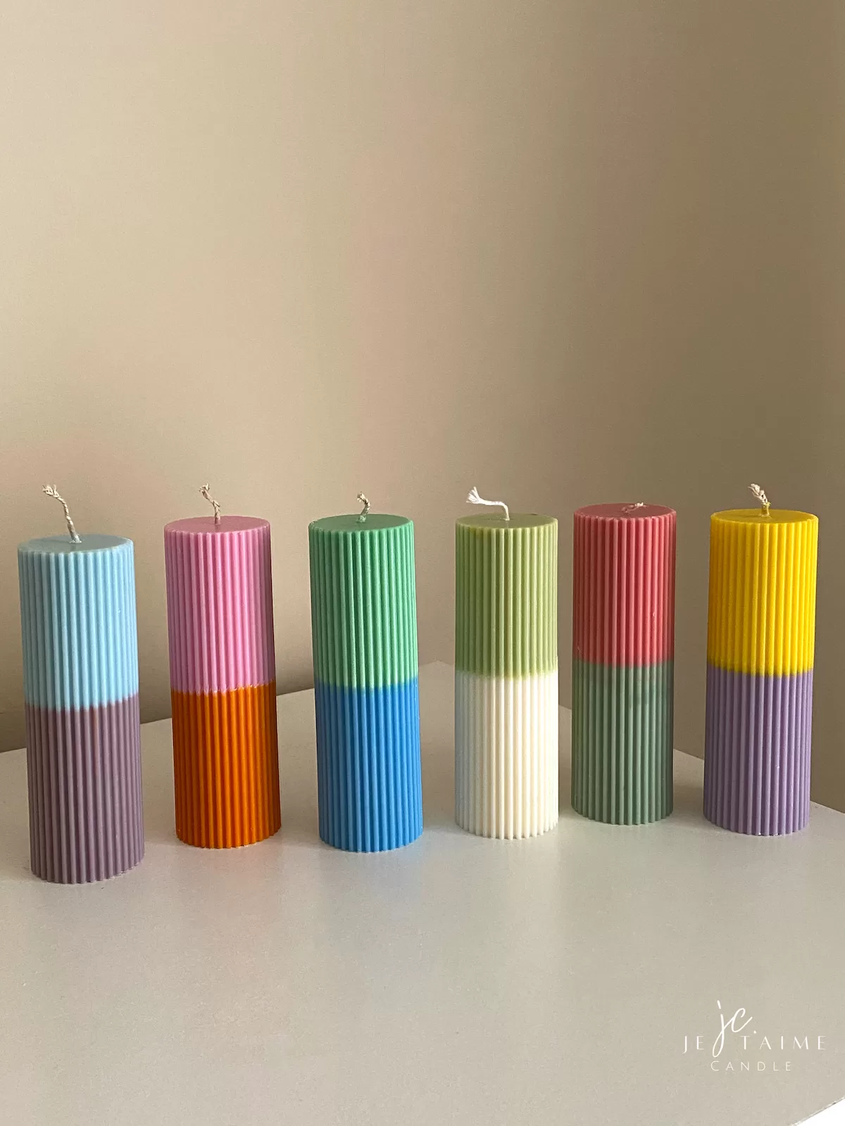 A row of tall ribbed pillar candles in pastel and bright two-tone colors, arranged on a white surface. The candles are displayed beside a green checkered lamp, a decorative mirror with an orange frame, and pampas grass in the background.
