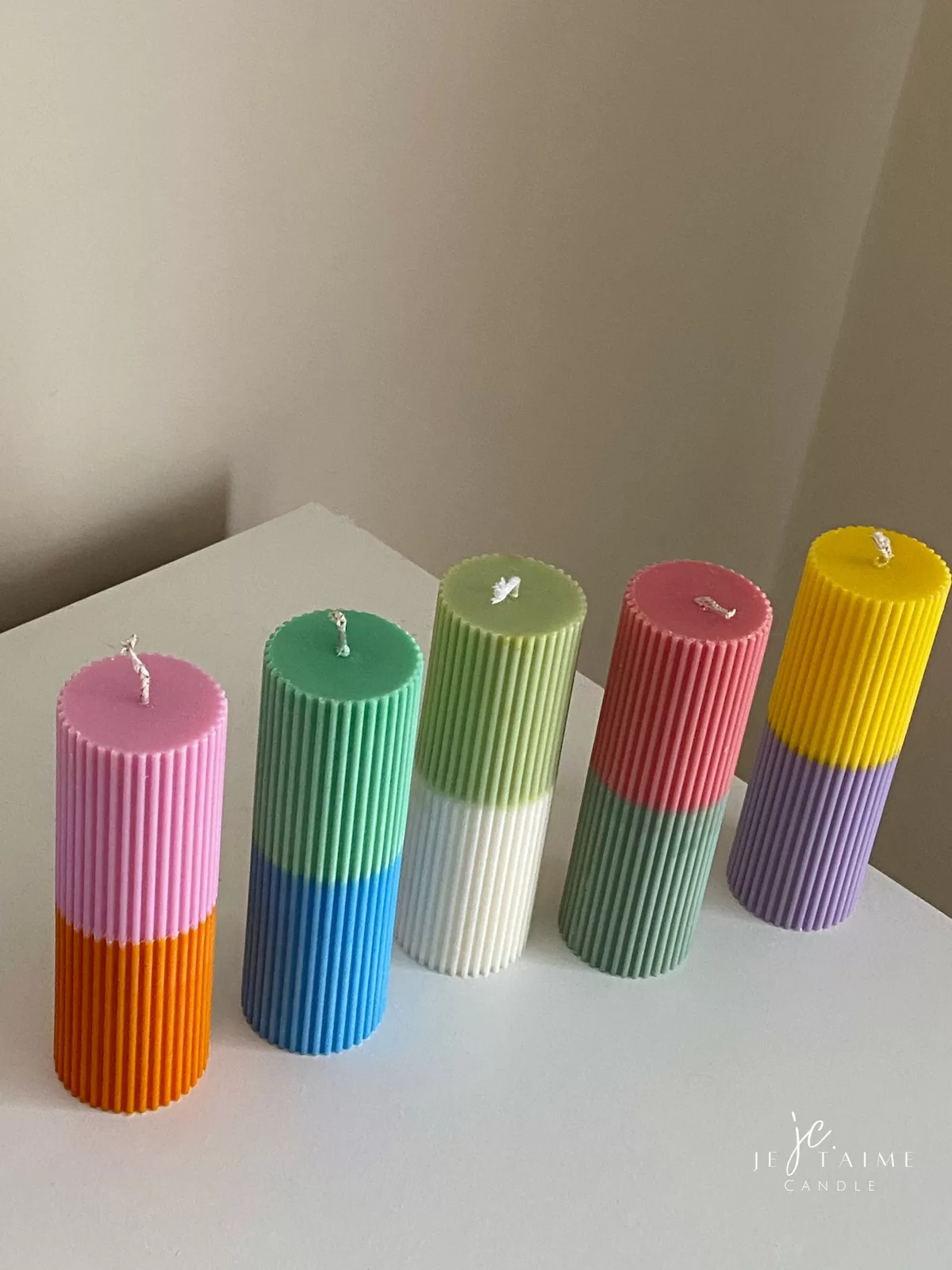 A row of tall ribbed pillar candles in pastel and bright two-tone colors, arranged on a white surface. The candles are displayed beside a green checkered lamp, a decorative mirror with an orange frame, and pampas grass in the background.
