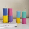 A row of tall ribbed pillar candles in pastel and bright two-tone colors, arranged on a white surface. The candles are displayed beside a green checkered lamp, a decorative mirror with an orange frame, and pampas grass in the background.