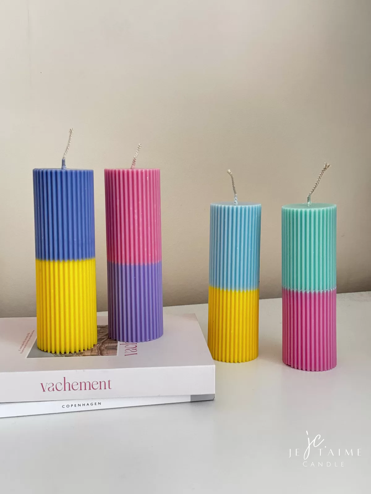 A row of tall ribbed pillar candles in pastel and bright two-tone colors, arranged on a white surface. The candles are displayed beside a green checkered lamp, a decorative mirror with an orange frame, and pampas grass in the background.