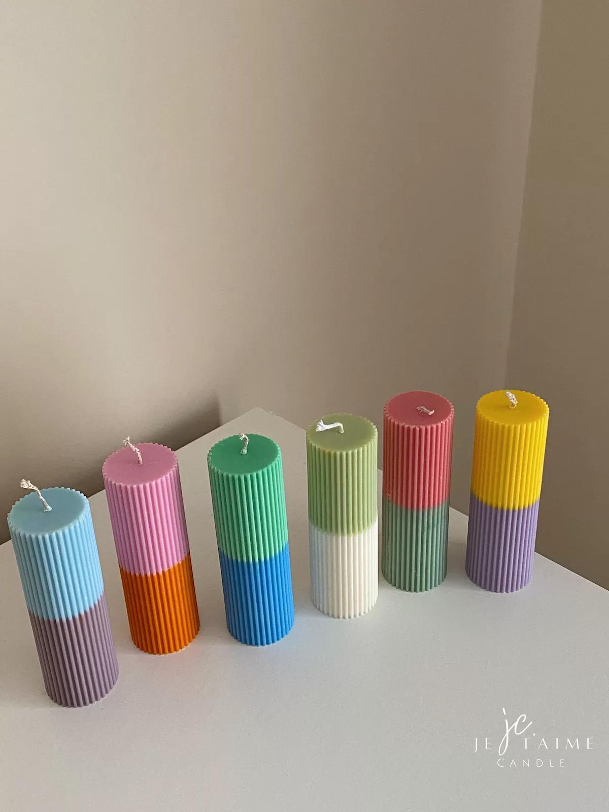 A row of tall ribbed pillar candles in pastel and bright two-tone colors, arranged on a white surface. The candles are displayed beside a green checkered lamp, a decorative mirror with an orange frame, and pampas grass in the background.