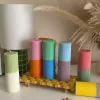 A row of tall ribbed pillar candles in pastel and bright two-tone colors, arranged on a white surface. The candles are displayed beside a green checkered lamp, a decorative mirror with an orange frame, and pampas grass in the background.