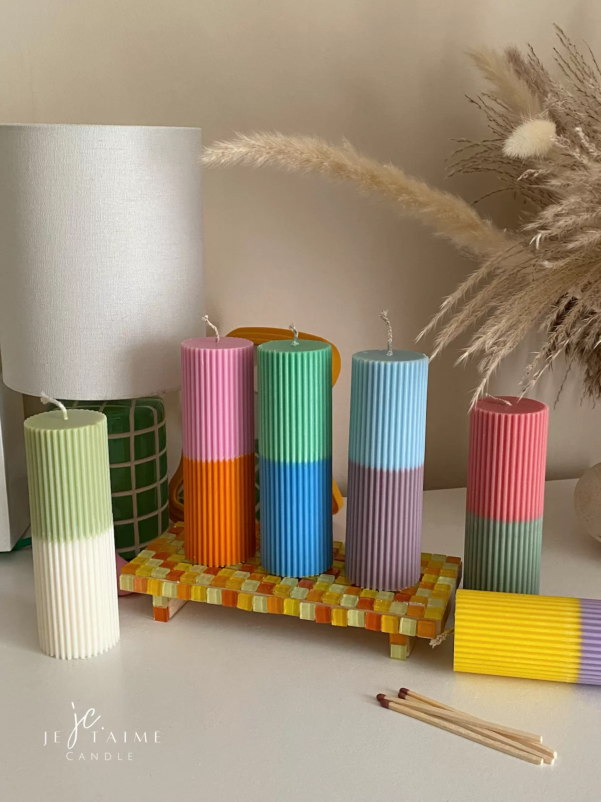 A row of tall ribbed pillar candles in pastel and bright two-tone colors, arranged on a white surface. The candles are displayed beside a green checkered lamp, a decorative mirror with an orange frame, and pampas grass in the background.