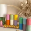 A row of tall ribbed pillar candles in pastel and bright two-tone colors, arranged on a white surface. The candles are displayed beside a green checkered lamp, a decorative mirror with an orange frame, and pampas grass in the background.