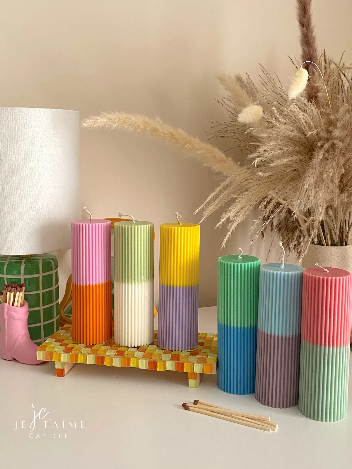 A row of tall ribbed pillar candles in pastel and bright two-tone colors, arranged on a white surface. The candles are displayed beside a green checkered lamp, a decorative mirror with an orange frame, and pampas grass in the background.