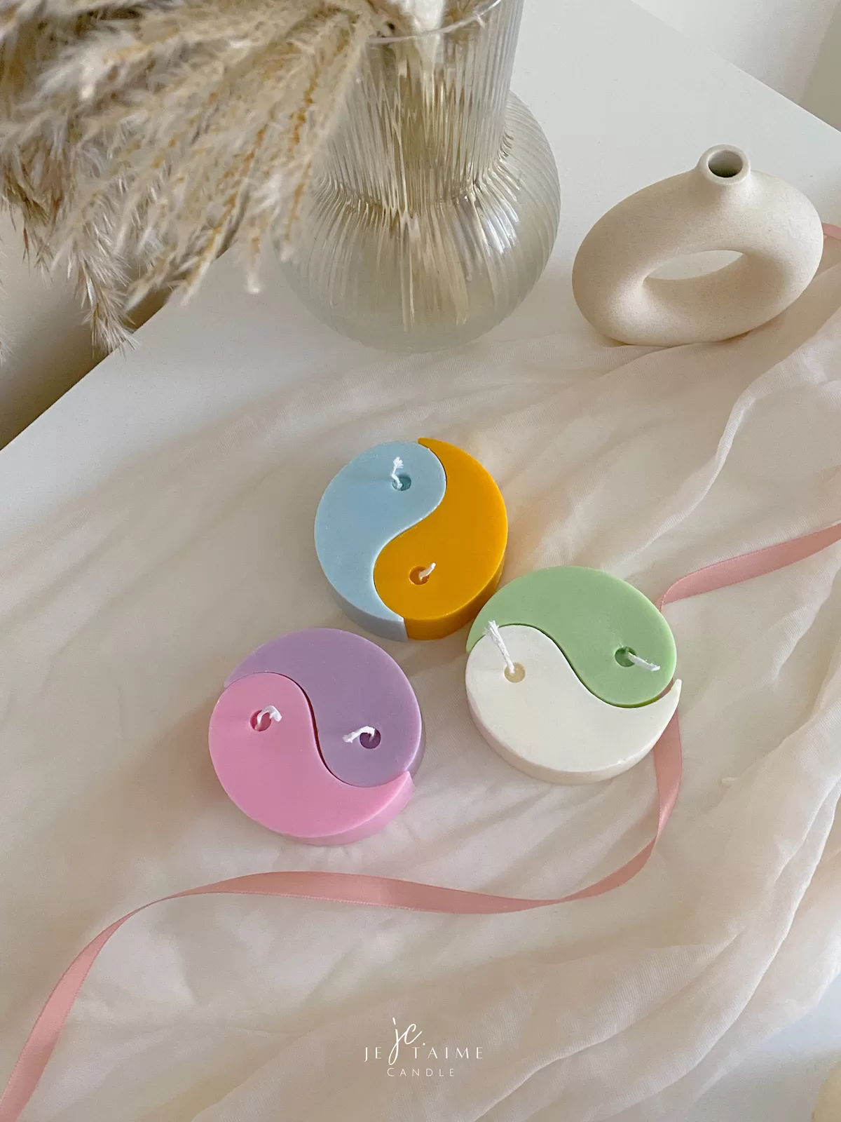 Four pastel yin-yang shaped candles in pairs of pink and purple, yellow and blue, green and cream, displayed on a white surface with a pink polka-dotted felt ball, a butter-themed box, and a disco ball nearby.