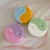 Four pastel yin-yang shaped candles in pairs of pink and purple, yellow and blue, green and cream, displayed on a white surface with a pink polka-dotted felt ball, a butter-themed box, and a disco ball nearby.