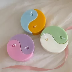 Four pastel yin-yang shaped candles in pairs of pink and purple, yellow and blue, green and cream, displayed on a white surface with a pink polka-dotted felt ball, a butter-themed box, and a disco ball nearby.