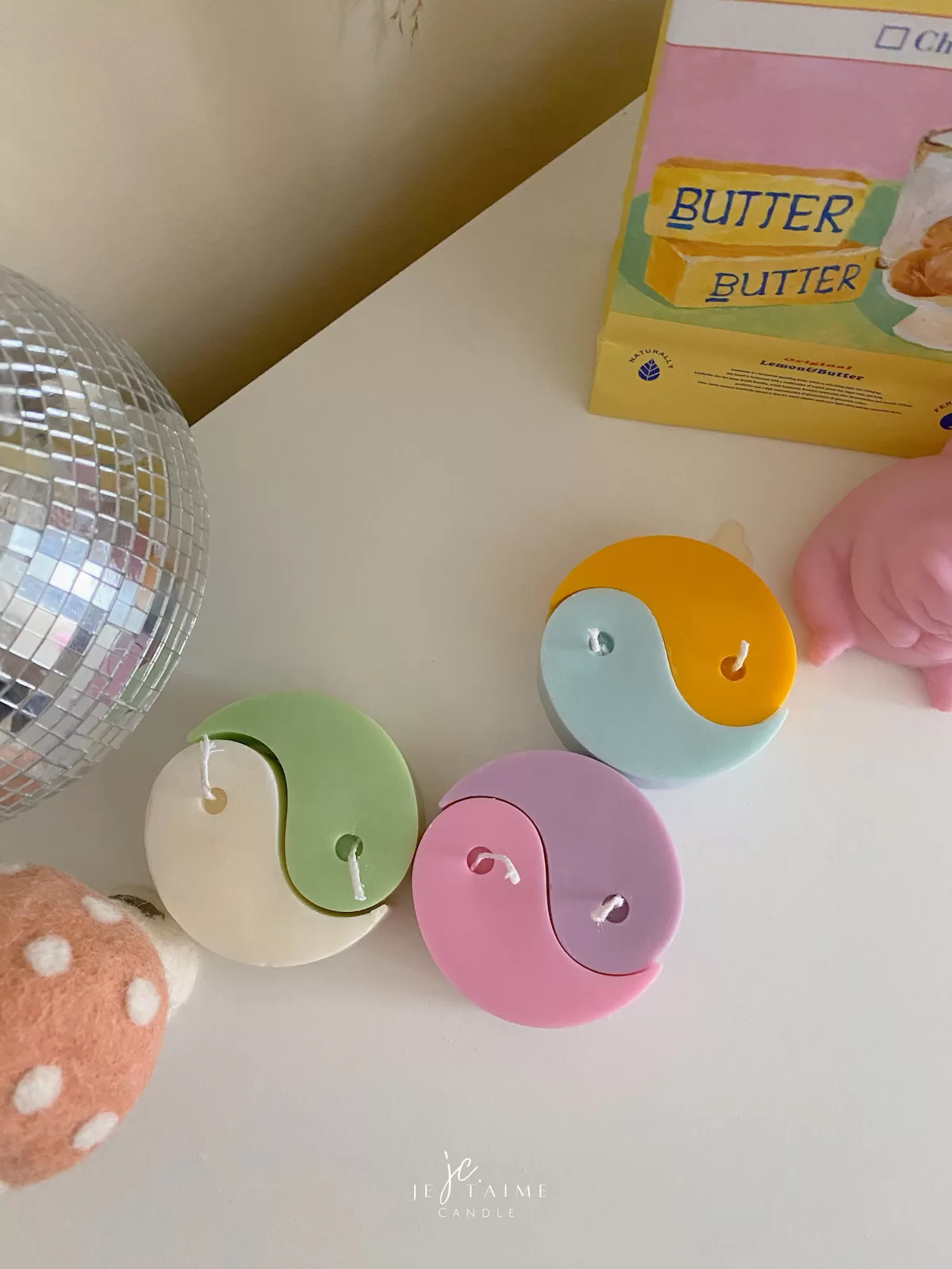 Four pastel yin-yang shaped candles in pairs of pink and purple, yellow and blue, green and cream, displayed on a white surface with a pink polka-dotted felt ball, a butter-themed box, and a disco ball nearby.