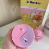 Four pastel yin-yang shaped candles in pairs of pink and purple, yellow and blue, green and cream, displayed on a white surface with a pink polka-dotted felt ball, a butter-themed box, and a disco ball nearby.