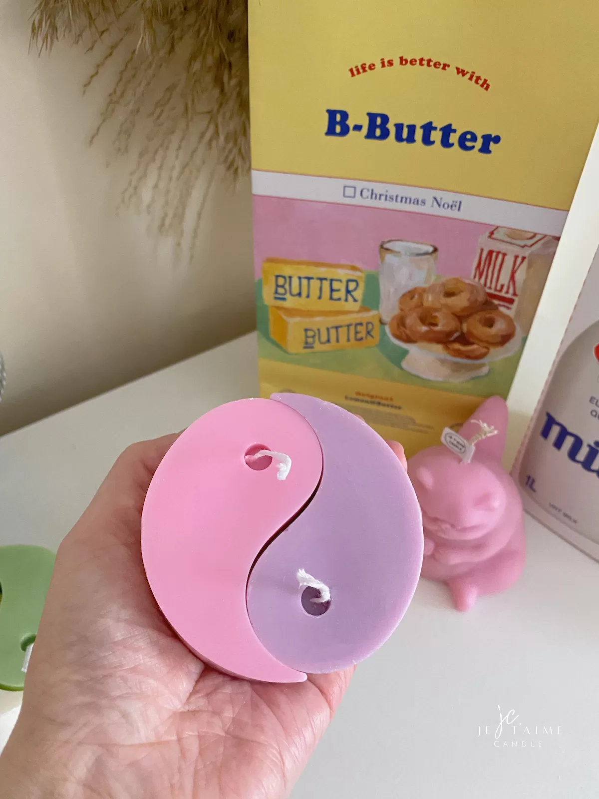 Four pastel yin-yang shaped candles in pairs of pink and purple, yellow and blue, green and cream, displayed on a white surface with a pink polka-dotted felt ball, a butter-themed box, and a disco ball nearby.