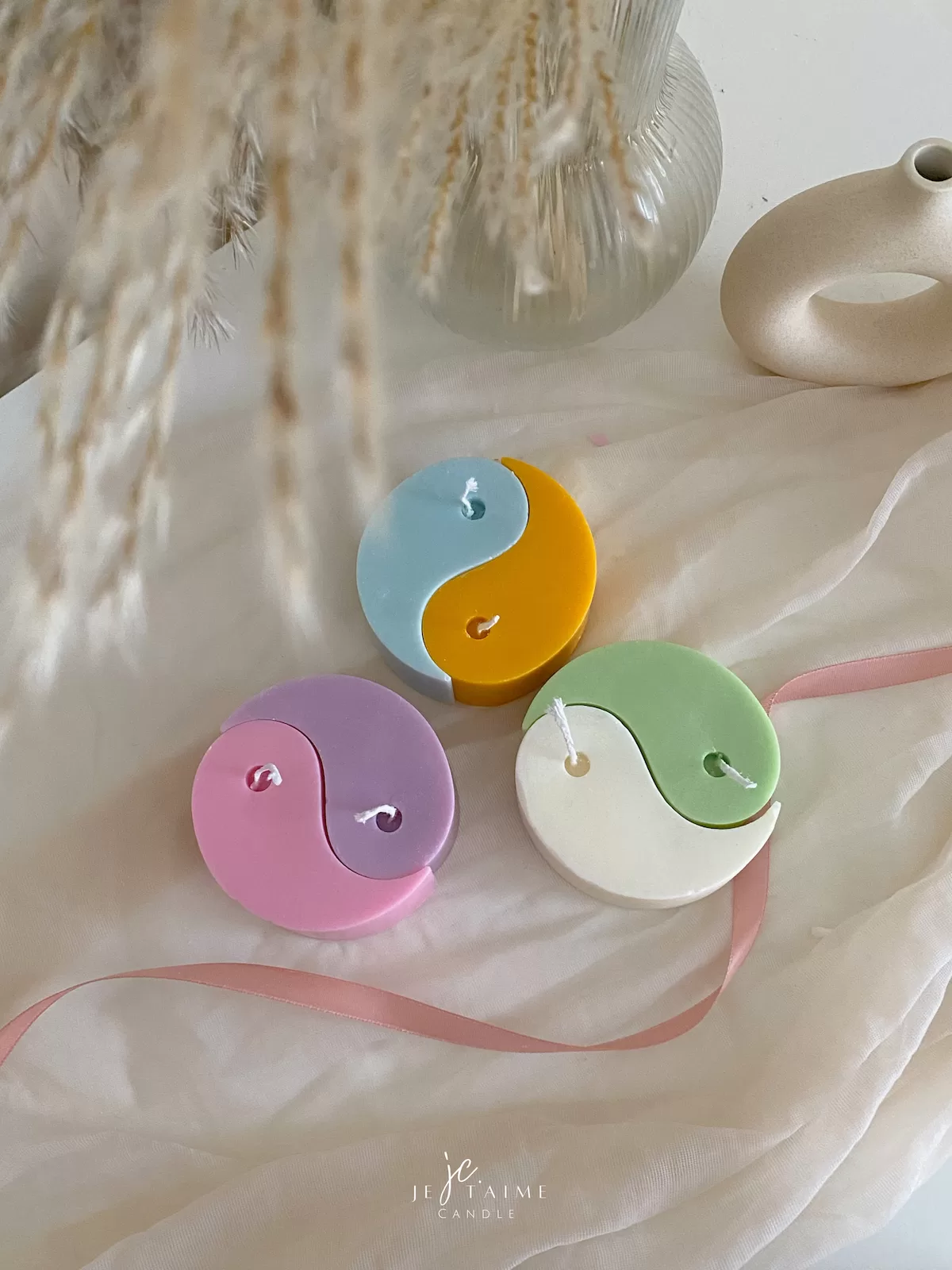 Four pastel yin-yang shaped candles in pairs of pink and purple, yellow and blue, green and cream, displayed on a white surface with a pink polka-dotted felt ball, a butter-themed box, and a disco ball nearby.