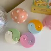 Four pastel yin-yang shaped candles in pairs of pink and purple, yellow and blue, green and cream, displayed on a white surface with a pink polka-dotted felt ball, a butter-themed box, and a disco ball nearby.