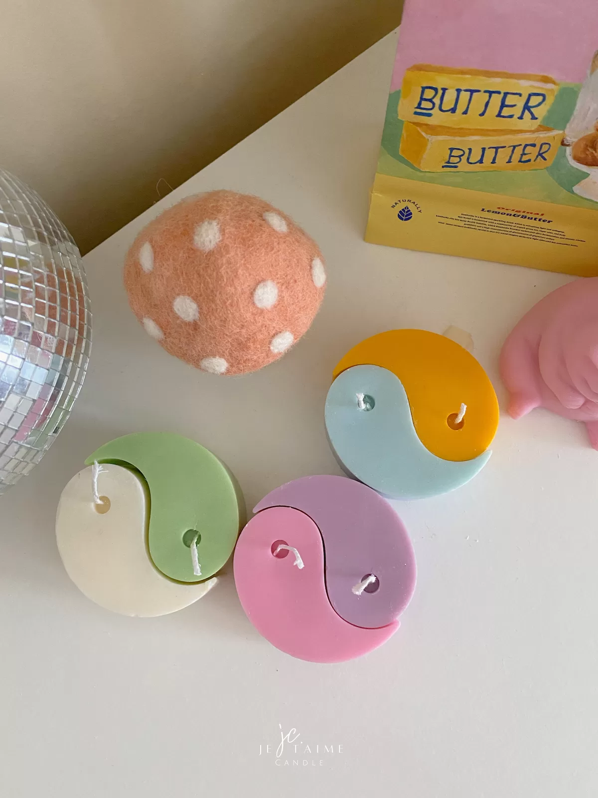 Four pastel yin-yang shaped candles in pairs of pink and purple, yellow and blue, green and cream, displayed on a white surface with a pink polka-dotted felt ball, a butter-themed box, and a disco ball nearby.