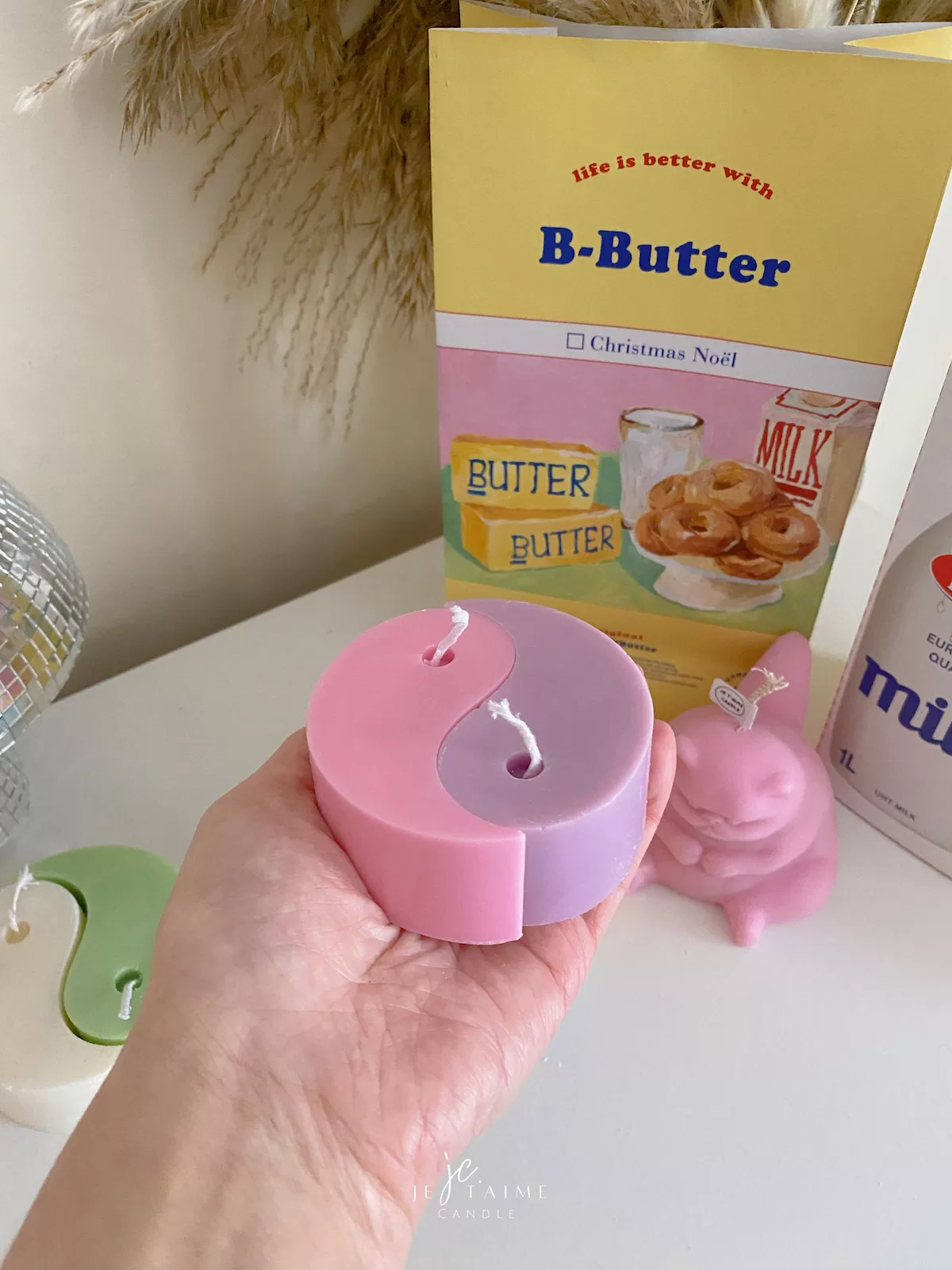 Four pastel yin-yang shaped candles in pairs of pink and purple, yellow and blue, green and cream, displayed on a white surface with a pink polka-dotted felt ball, a butter-themed box, and a disco ball nearby.