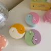 Four pastel yin-yang shaped candles in pairs of pink and purple, yellow and blue, green and cream, displayed on a white surface with a pink polka-dotted felt ball, a butter-themed box, and a disco ball nearby.