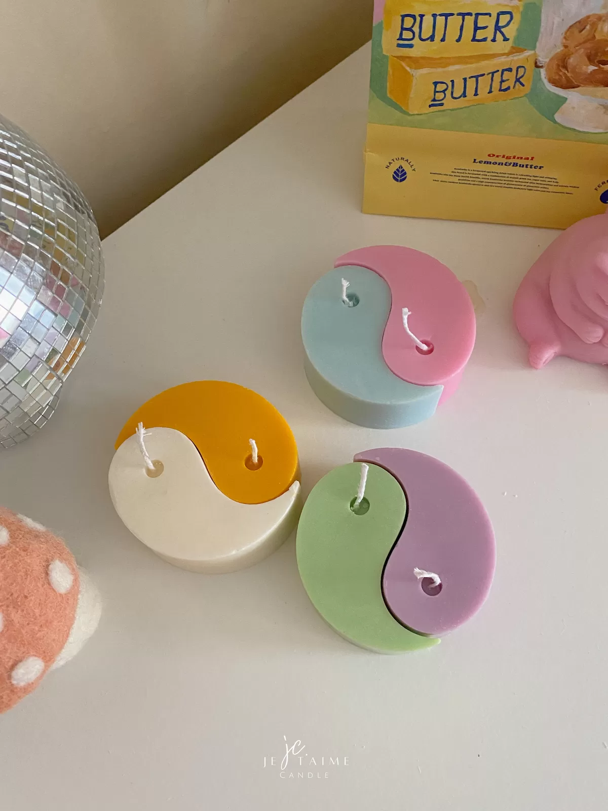 Four pastel yin-yang shaped candles in pairs of pink and purple, yellow and blue, green and cream, displayed on a white surface with a pink polka-dotted felt ball, a butter-themed box, and a disco ball nearby.