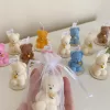Colorful teddy bear candles in pink, blue, brown, orange, and white, wrapped in white organza bags with ribbons and personalized tags, displayed on a green tiled box with pampas grass decor in the background