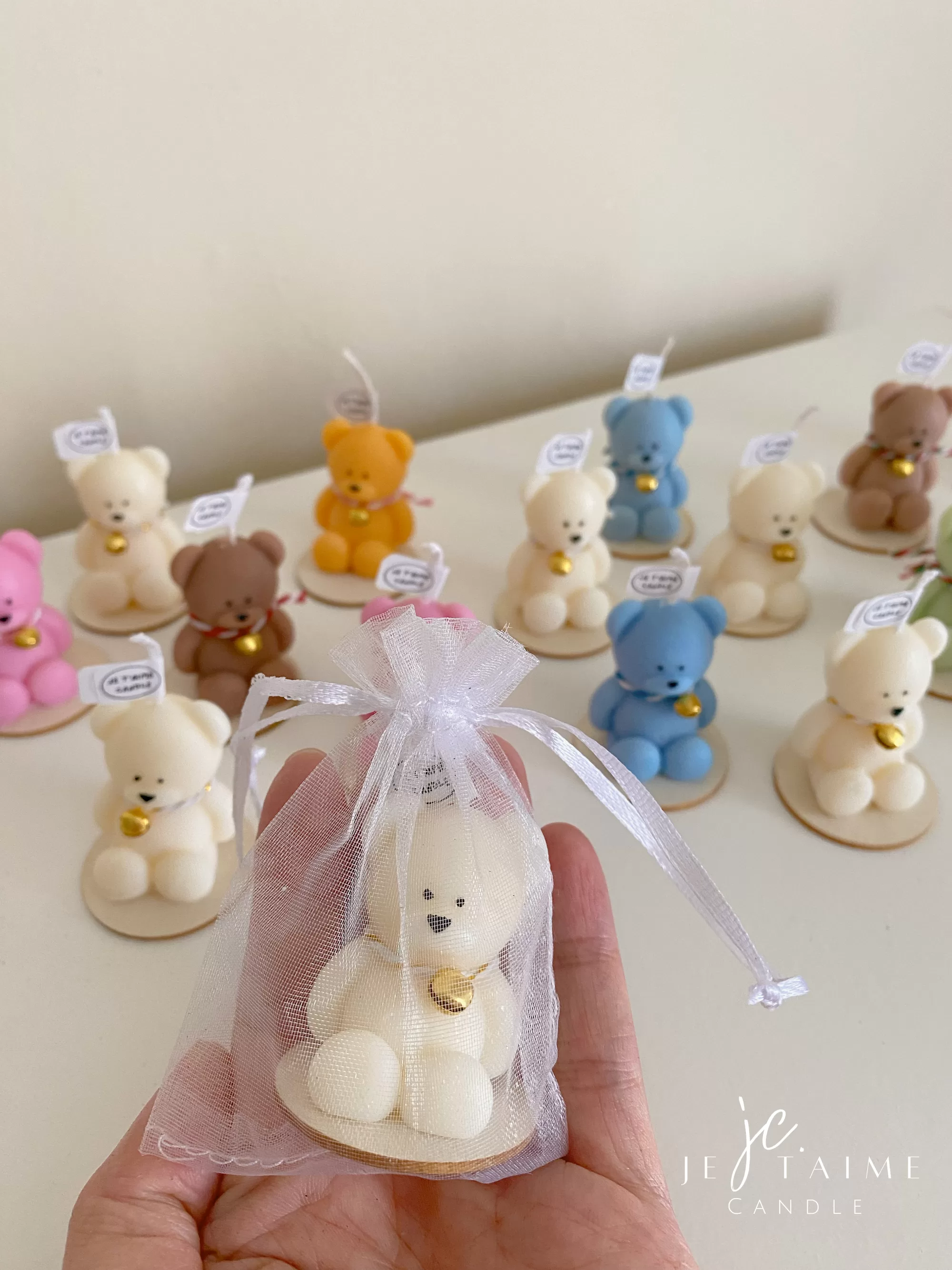 Colorful teddy bear candles in pink, blue, brown, orange, and white, wrapped in white organza bags with ribbons and personalized tags, displayed on a green tiled box with pampas grass decor in the background