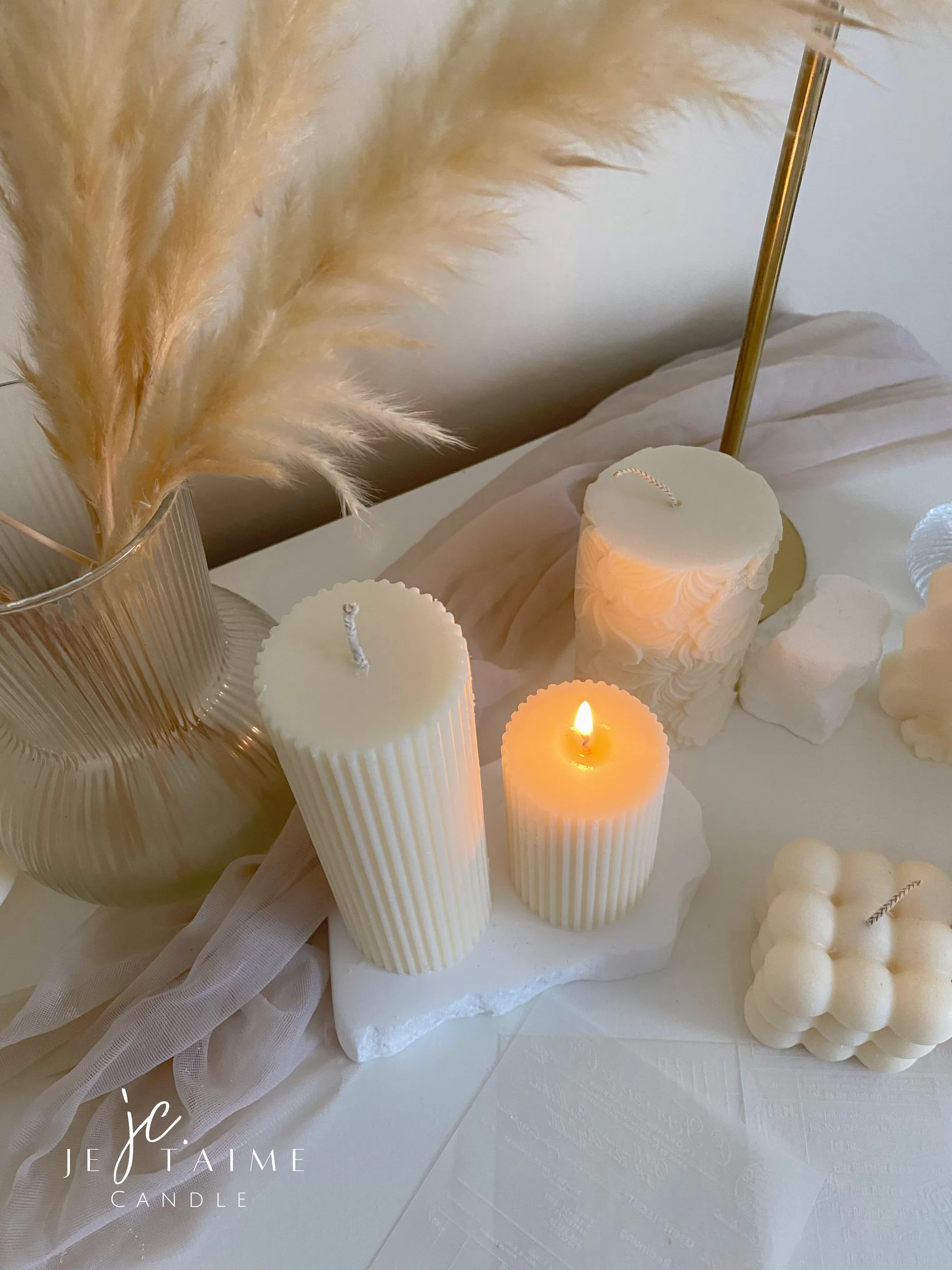 Two handmade ribbed pillar candles in ivory white soy wax, one tall and one short, placed on a white stone base beside pampas grass, a gold candle holder, and decorative candles. The ribbed texture gives these candles a modern minimalist look, making them ideal for wedding centerpieces, cozy home décor, or as stylish handmade gifts. Their clean design and neutral color make them versatile decorative accents that fit perfectly into Scandinavian, modern, and rustic interiors.