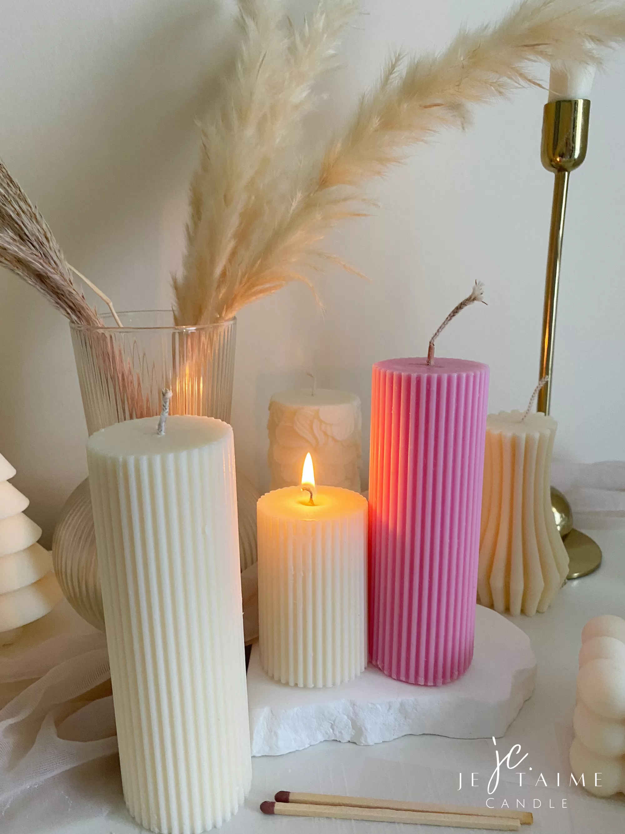 Two handmade ribbed pillar candles in ivory white and pink soy wax, one tall and one short, placed on a white stone base beside pampas grass, a gold candle holder, and decorative candles. The ribbed texture gives these candles a modern minimalist look, making them ideal for wedding centerpieces, cozy home décor, or as stylish handmade gifts. Their clean design and neutral color make them versatile decorative accents that fit perfectly into Scandinavian, modern, and rustic interiors.