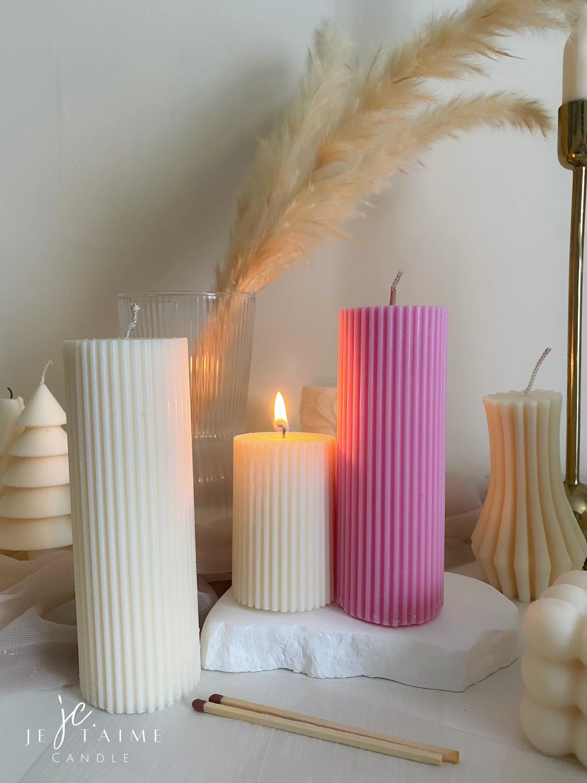 Two handmade ribbed pillar candles in ivory white and pink soy wax, one tall and one short, placed on a white stone base beside pampas grass, a gold candle holder, and decorative candles. The ribbed texture gives these candles a modern minimalist look, making them ideal for wedding centerpieces, cozy home décor, or as stylish handmade gifts. Their clean design and neutral color make them versatile decorative accents that fit perfectly into Scandinavian, modern, and rustic interiors.