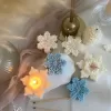 A collection of decorative candles in various shapes, including snowflakes, a Christmas tree, a nutcracker, a house, and a cylindrical candle. The snowflake candles are intricately designed in white and blue, displayed on soft fabric.