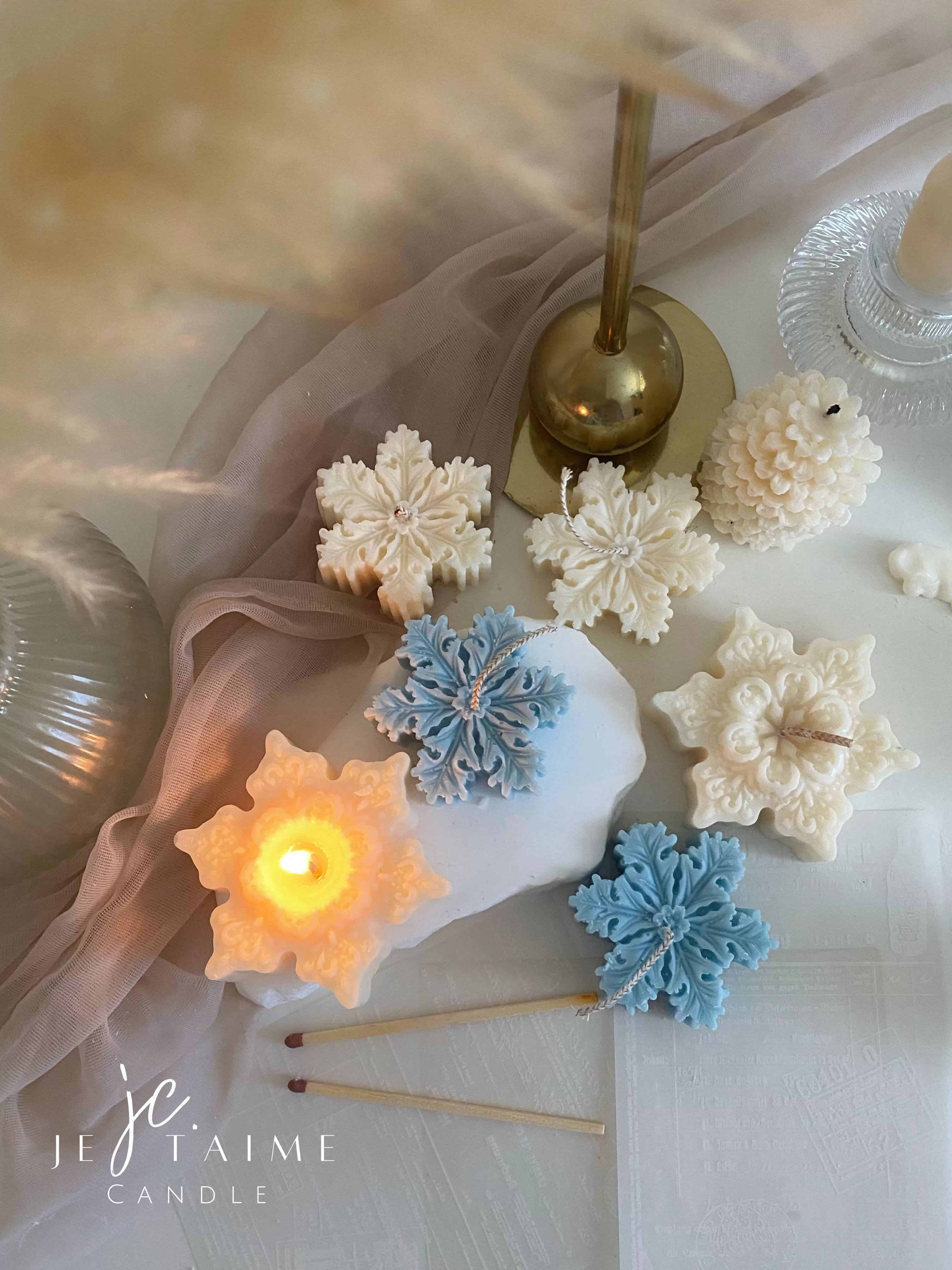 A collection of decorative candles in various shapes, including snowflakes, a Christmas tree, a nutcracker, a house, and a cylindrical candle. The snowflake candles are intricately designed in white and blue, displayed on soft fabric.