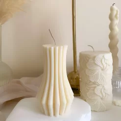 Handmade vase column aesthetic candle in ivory soy wax with a modern fluted design, displayed on a white stone base with pampas grass and other decorative candles in the background. This sculptural column candle is perfect for minimalist home décor, wedding centerpieces, or as a unique handmade gift, adding timeless elegance and modern style to any space.