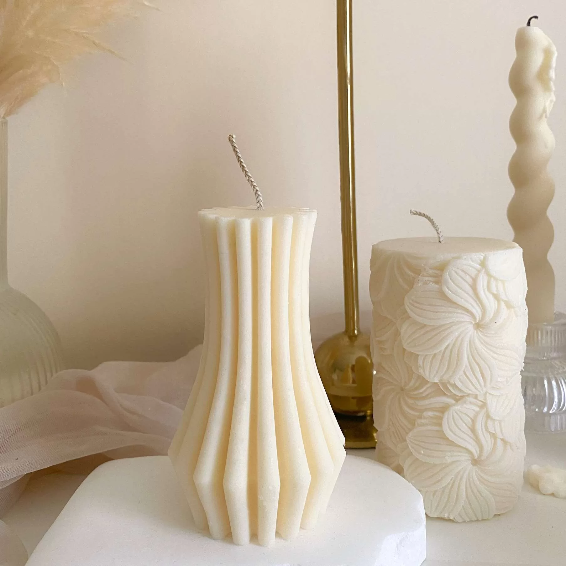 Handmade vase column aesthetic candle in ivory soy wax with a modern fluted design, displayed on a white stone base with pampas grass and other decorative candles in the background. This sculptural column candle is perfect for minimalist home décor, wedding centerpieces, or as a unique handmade gift, adding timeless elegance and modern style to any space.
