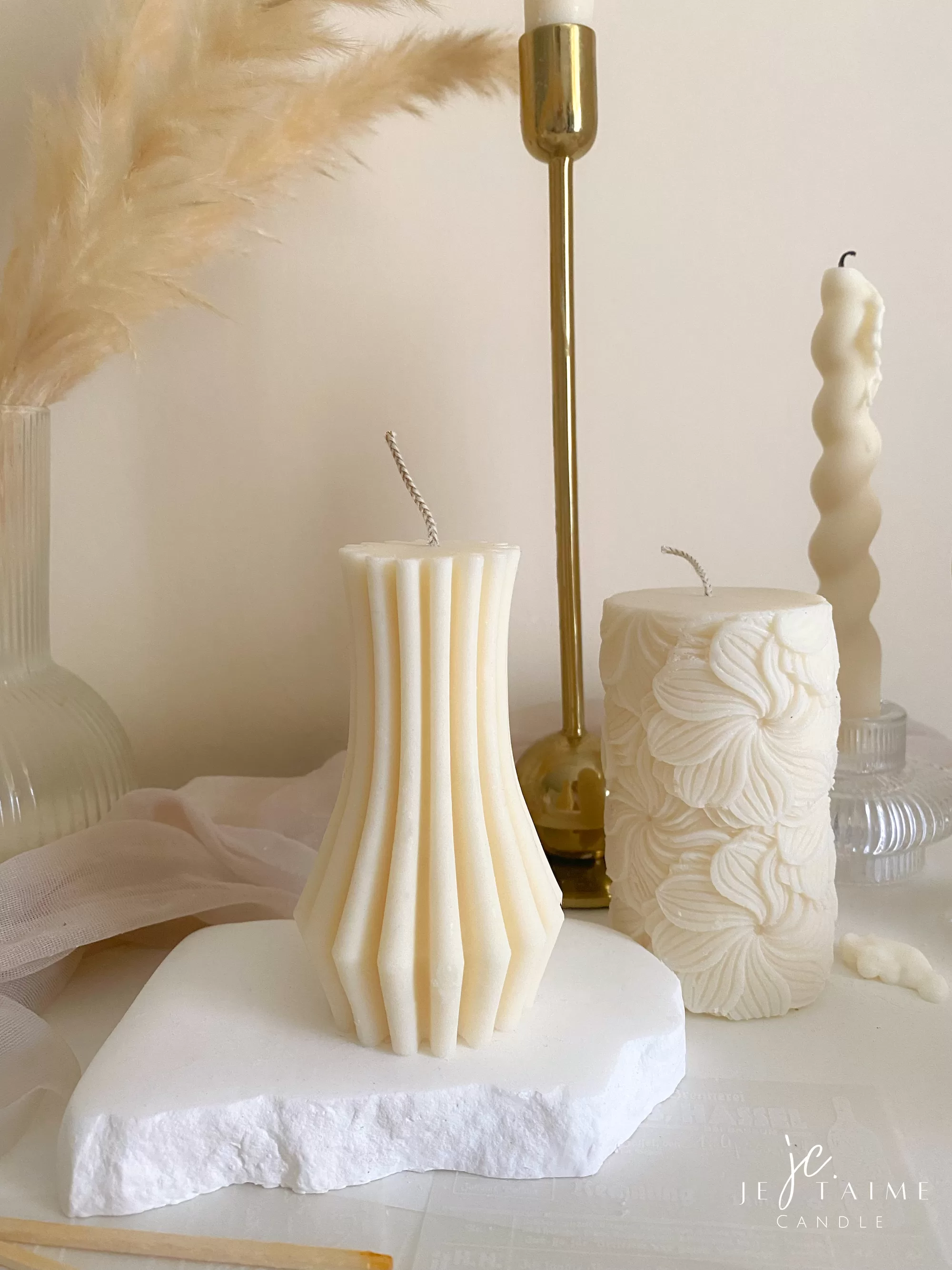 Handmade vase column aesthetic candle in ivory soy wax with a modern fluted design, displayed on a white stone base with pampas grass and other decorative candles in the background. This sculptural column candle is perfect for minimalist home décor, wedding centerpieces, or as a unique handmade gift, adding timeless elegance and modern style to any space.