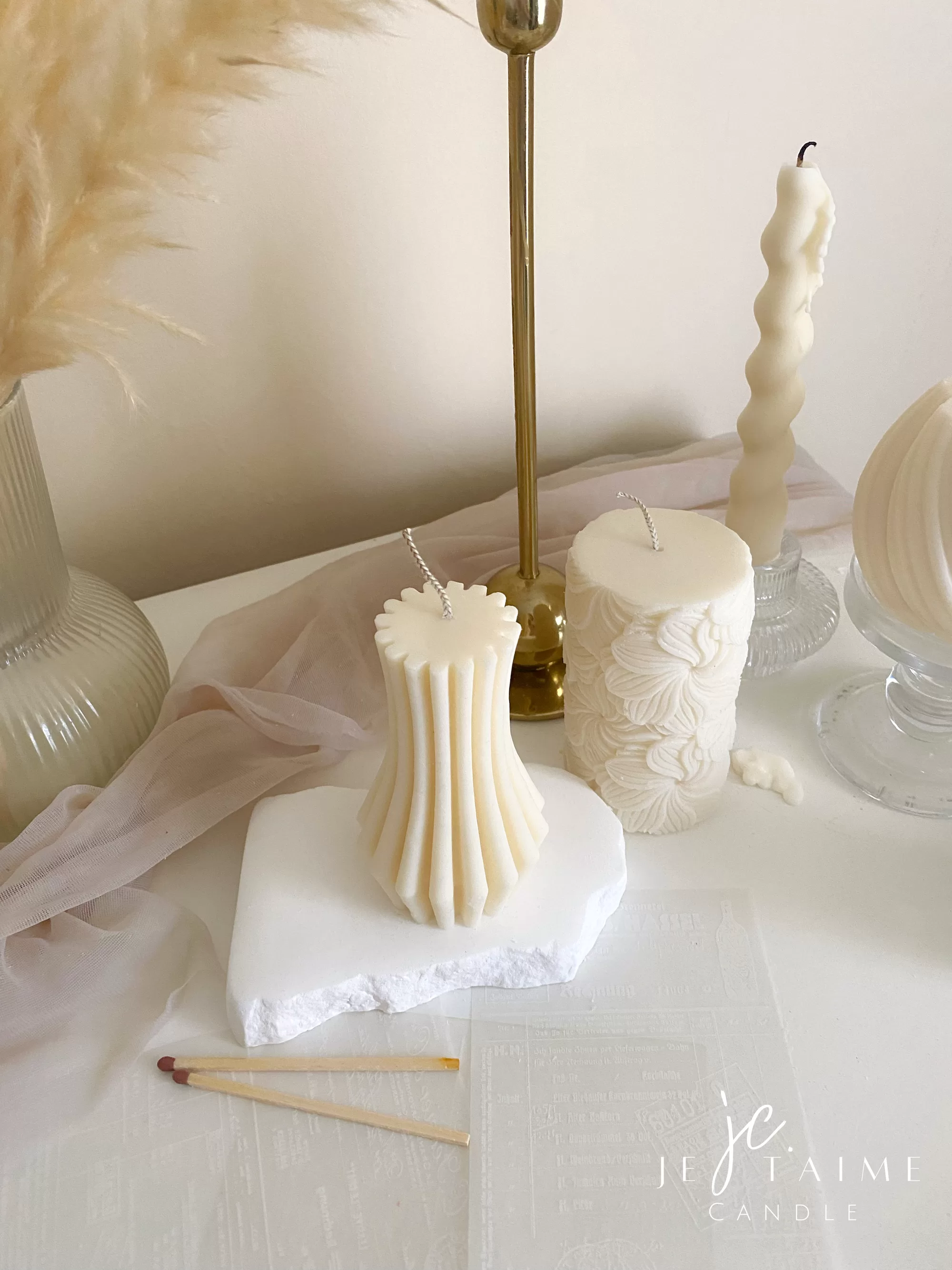 Handmade vase column aesthetic candle in ivory soy wax with a modern fluted design, displayed on a white stone base with pampas grass and other decorative candles in the background. This sculptural column candle is perfect for minimalist home décor, wedding centerpieces, or as a unique handmade gift, adding timeless elegance and modern style to any space.