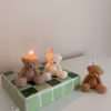 The image shows a group of teddy bear-shaped candles arranged neatly in rows on a light surface. Each bear candle has a wick on top of its head. The texture and detailing make them look fluffy and realistic. In the lower-left corner, there’s a logo that says 