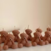 The image shows a group of teddy bear-shaped candles arranged neatly in rows on a light surface. Each bear candle has a wick on top of its head. The texture and detailing make them look fluffy and realistic. In the lower-left corner, there’s a logo that says 