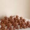The image shows a group of teddy bear-shaped candles arranged neatly in rows on a light surface. Each bear candle has a wick on top of its head. The texture and detailing make them look fluffy and realistic. In the lower-left corner, there’s a logo that says 