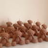 The image shows a group of teddy bear-shaped candles arranged neatly in rows on a light surface. Each bear candle has a wick on top of its head. The texture and detailing make them look fluffy and realistic. In the lower-left corner, there’s a logo that says 