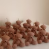 The image shows a group of teddy bear-shaped candles arranged neatly in rows on a light surface. Each bear candle has a wick on top of its head. The texture and detailing make them look fluffy and realistic. In the lower-left corner, there’s a logo that says 
