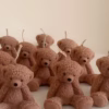 The image shows a group of teddy bear-shaped candles arranged neatly in rows on a light surface. Each bear candle has a wick on top of its head. The texture and detailing make them look fluffy and realistic. In the lower-left corner, there’s a logo that says 