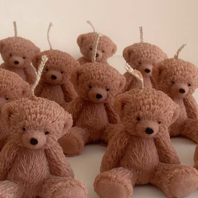 The image shows a group of teddy bear-shaped candles arranged neatly in rows on a light surface. Each bear candle has a wick on top of its head. The texture and detailing make them look fluffy and realistic. In the lower-left corner, there’s a logo that says "JE T'AIME CANDLE"