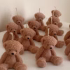 The image shows a group of teddy bear-shaped candles arranged neatly in rows on a light surface. Each bear candle has a wick on top of its head. The texture and detailing make them look fluffy and realistic. In the lower-left corner, there’s a logo that says 