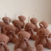 The image shows a group of teddy bear-shaped candles arranged neatly in rows on a light surface. Each bear candle has a wick on top of its head. The texture and detailing make them look fluffy and realistic. In the lower-left corner, there’s a logo that says 