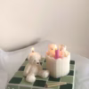 The image shows a group of teddy bear-shaped candles arranged neatly in rows on a light surface. Each bear candle has a wick on top of its head. The texture and detailing make them look fluffy and realistic. In the lower-left corner, there’s a logo that says 