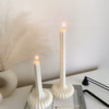 “Two lit sculptural candles with ribbed bases placed on top of a stack of design books, beside a candle snuffer and decorative pampas grass in a minimal, modern setting.