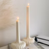 “Two lit sculptural candles with ribbed bases placed on top of a stack of design books, beside a candle snuffer and decorative pampas grass in a minimal, modern setting.