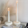 Two lit sculptural candles with ribbed bases placed on top of a stack of design books, beside a candle snuffer and decorative pampas grass in a minimal, modern setting.