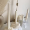 “Two lit sculptural candles with ribbed bases placed on top of a stack of design books, beside a candle snuffer and decorative pampas grass in a minimal, modern setting.