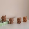 Three bear-shaped candles are placed on a white surface. Two of the candles are lit, with flames on top of their heads. The candles have round heads, small ears, and simple facial features. One candle is positioned on a green-tiled block, while the other two stand directly on the surface.