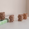 Three bear-shaped candles are placed on a white surface. Two of the candles are lit, with flames on top of their heads. The candles have round heads, small ears, and simple facial features. One candle is positioned on a green-tiled block, while the other two stand directly on the surface.